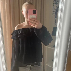 Svart offshoulder blus - Säljer en elegant svart offshoulder blus med volangdetaljer. Blusen har långa ärmar och en luftig passform, perfekt för en stilren look.