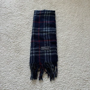 Rutig halsduk från Burberry - Säljer en klassisk rutig halsduk från Burberry i mörkblått med inslag av rött och grått. Tillverkad av 100% lammull, vilket gör den både mjuk och varm. Perfekt accessoar för kyliga dagar. Fransen ger en extra touch av stil.