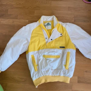 Gul och vit vindjacka - Säljer en retro gul och vit vindjacka/skidjacka.  Jackan har en dragkedja och knappar framtill samt praktiska fickor. Köpt second hand men aldrig använt. 