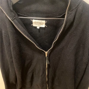 Svart stickad tröja med dragkedja - Säljer en svart stickad tröja från Maison Margiela med dragkedja framtill. Tröjan har en hög krage och långa ärmar, perfekt för kyligare dagar. Den är stilren och enkel att matcha med olika outfits.