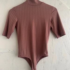 Ribbad  bodysuit NA-KD - Snygg ribbad bodysuit i en mjuk brunrosa nyans. Den har korta ärmar och en hög krage som ger en stilren look. Perfekt för att matcha med jeans eller kjol för en trendig outfit.