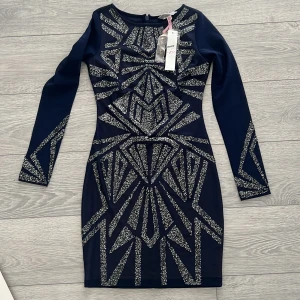 Mörkblå glittrig klänning från lipsy - Snygg mörkblå klänning från lipsy med glittrigt geometriskt mönster. Den har en tight passform och långa ärmar, perfekt för en festlig kväll. Klänningen är tillverkad i ett syntetmaterial och har en dragkedja i ryggen.