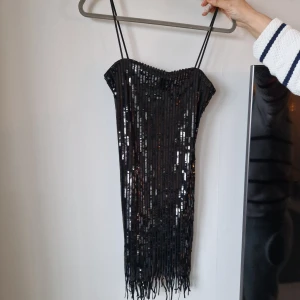 Svart paljettklänning med fransar - Snygg svart klänning med paljetter och fransar. Axelbandslös design med smala axelband. Perfekt för en festlig kväll!
