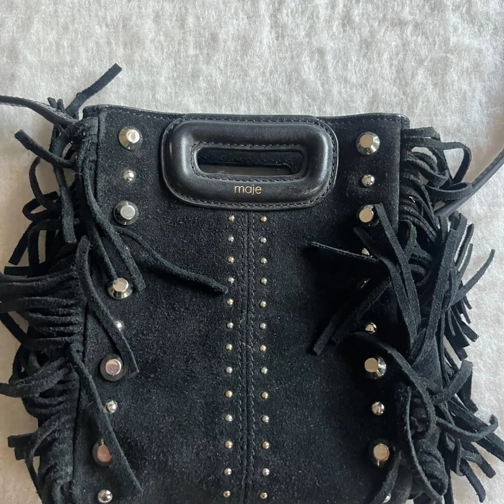 Snygg svart axelväska i mocka från Maje med coola fransdetaljer och nitar. Väskan har ett praktiskt handtag och en stilren design som passar perfekt för att ge din outfit en edgy touch. Perfekt för när man ska iväg men inte vill ha stor väska då den ändå rymmer mycket🖤strlk mini! Nypris ca 2500. Laukut & Käsilaukut.