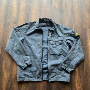 Vindjacka/overshirt från Stone Island - Säljer en stilren navy jacka/overshirt från Stone Island med dragkedja och en knappförsedd bröstficka. Jackan har långa ärmar och en klassisk krage. Perfekt för en avslappnad look. Priset kan diskutera, hör av dig vis minsta fundering!