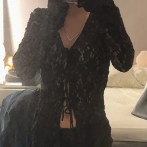 Svart spetsblus med knytning - Säljer en elegant svart spetsblus med knytning framtill. Blusen har långa ärmar och en djup v-ringning som ger en stilren look. Perfekt för att klä upp en outfit med en touch av elegans. Passar xxs/M
