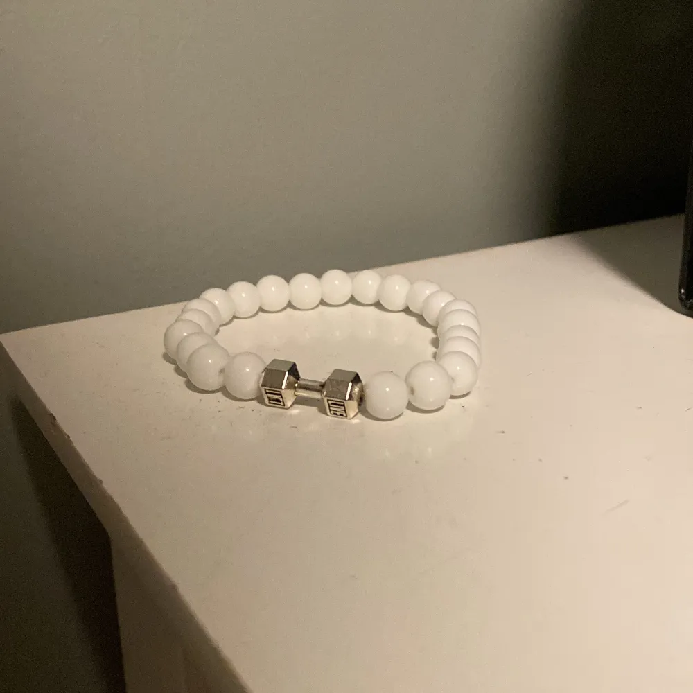 Snyggt vitt armband med pärlor och en unik silverdetalj i form av en hantel. Perfekt accessoar för att ge din outfit en sportig touch.. Asusteet.