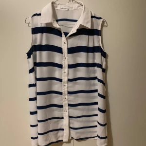 Randig ärmlös skjorta - Säljer en stilren ärmlös skjorta med breda blå och vita ränder. Skjortan har knappar framtill och en klassisk krage. Perfekt för en avslappnad och trendig look.