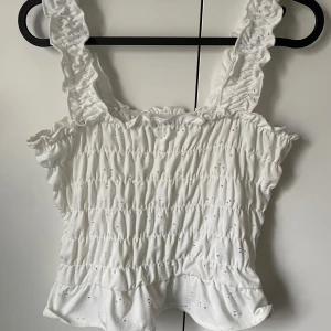 Vit ärmlös topp med volangdetaljer - Säljer en söt vit ärmlös topp med volangdetaljer och smockad design. Toppen har breda axelband och en luftig passform. Storlek M men passar även S samt Xs. 
