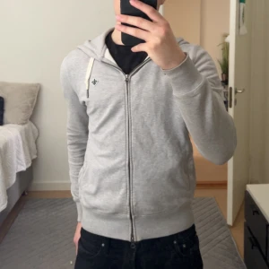 Morris hoodie - Säljer en stilren grå Morris hoodie med dragkedja och ribbade muddar. Perfekt för en avslappnad look. Den har en liten logga på bröstet och en bekväm passform. Enda defekten är att fickan inuti hoodien är sönder men det är inget som syns och kan lätt sys.