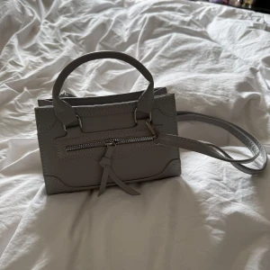 Grå axelväska med dragkedja - Snygg grå axelväska med praktisk dragkedja framtill. Väskan har både handtag och en avtagbar, justerbar axelrem. Väskan är 20 cm bred och 13 cm hög. Oanvänd