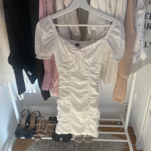 Vit klänning med puffärmar - Säljer en söt vit klänning med puffiga kortärmar och v-ringad design. Klänningen har ett fint broderat mönster och rynkade detaljer som ger en smickrande passform. Perfekt för sommarens festligheter!