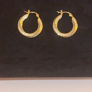 Guldiga örhängen med stenar Celine  - Snygga guldiga hoop-örhängen dekorerade med glittrande stenar. Perfekta för att ge en touch av glamour till din outfit. Lätta att bära och passar till många olika stilar.