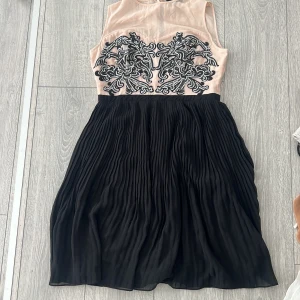 Svart och beige klänning från Spotlight by Warehouse - Elegant klänning med en beige överdel och svart kjol. Överdelen har ett detaljerat svart broderi och är ärmlös. Kjolen är plisserad och ger en fin rörelse. Perfekt för speciella tillfällen. Insydd på axlarna använd en gång 