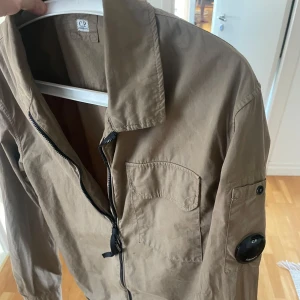 Beige overshirt från CP Company - Säljer en beige jacka från CP Company med dragkedja och en ficka på bröstet. Jackan har en cool detalj på ärmen och är perfekt för vår och höst. Den har långa ärmar och en klassisk krage storlek M och sitter perfekt kom pm för mer info .