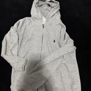 Grå hoodie från Polo Ralph Lauren - Säljer en klassisk grå hoodie från Polo Ralph Lauren med dragkedja och huva. Den har en liten broderad logotyp på bröstet och är tillverkad i mjukt material för extra komfort. Priset går att diskutera vid snabb köp.