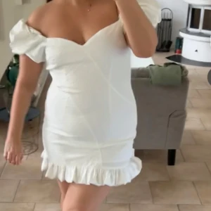 Vit offshoulder klänning med volang - Säljer en söt vit offshoulder klänning med volangdetaljer vid fållen. Klänningen har en tight passform och är perfekt för sommarens festligheter. Den har en v-ringad design och kort längd som ger en chic look.