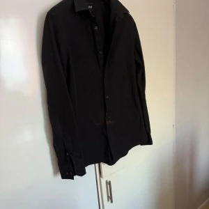 H&M - Snygg svart skjorta från H&M med långa ärmar och knappar framtill. Perfekt för en stilren look. Skjortan är i ett lättstruket material vilket gör den enkel att underhålla.