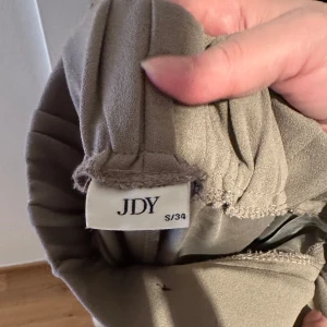 Beige kostymbyxor från JDY - Snygga beige kostymbyxor från JDY med hög midja och vida ben. Byxorna har två stora knappar framtill och veckade detaljer som ger en elegant look. Perfekta för en stilren outfit.