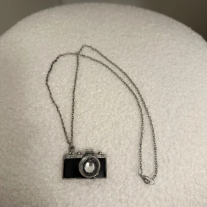 Halsband med kamerahänge - Unikt halsband med ett kamerahänge i silver och svart. Perfekt för fotointresserade! Kedjan är tunn och smidig med ett klassiskt lås.