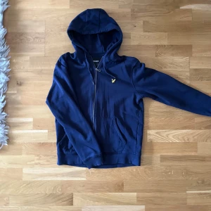 Blå hoodie från Lyle & Scott - Säljer en snygg blå hoodie från Lyle & Scott med dragkedja och logga på bröstet. Tröjan har en  huva med snörning och en ficka framtill. Perfekt för en avslappnad stil. Pris kan diskuteras!