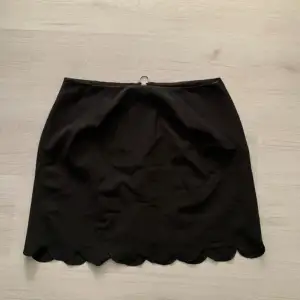 Snygg svart kortkjol med en dekorativ dragkedja baktill och vågig kant nedtill. Perfekt för en stilren look.