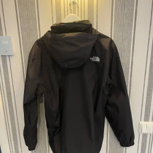 Svart vindjacka från The North Face - Säljer en stilren svart vindjacka från The North Face. Jackan har en praktisk huva och långa ärmar. Perfekt för blåsiga dagar och har en diskret logga på bröstet. Passar bra för utomhusaktiviteter.