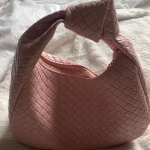 Rosa axelväska med knyt - Snygg rosa axelväska med ett flätat mönster och en praktisk dragkedja. Väskan har en unik knytning på handtaget som ger den en trendig look. Perfekt för att ge din outfit en färgklick!