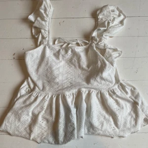 Vit spets blus med volangdetaljer - Charmig vit spets blus med volangdetaljer och knytband i ryggen. Blusen har ett vackert broderat mönster och är ärmlös med breda axelband. Perfekt för en somrig look! Pris kan diskuteras vid snabb affär!