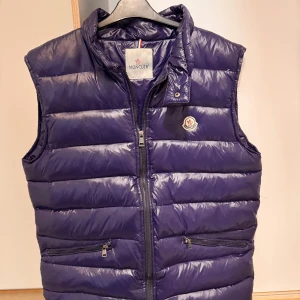 Mörkblå dunväst från Moncler - Snygg mörkblå dunväst från Moncler med dragkedja och två praktiska fickor framtill. Västen har en quiltad design och Monclers logotyp på bröstet. Perfekt för kyligare dagar när du vill hålla stilen.