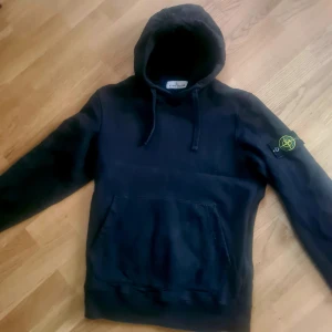 Svart hoodie från Stone Island - Säljer en svart hoodie från Stone Island med klassisk logga på ärmen. Tröjan har en stor ficka framtill och justerbar huva med dragsko. Perfekt för en stilren och avslappnad look....stl 170,,passar i vuxen xs 