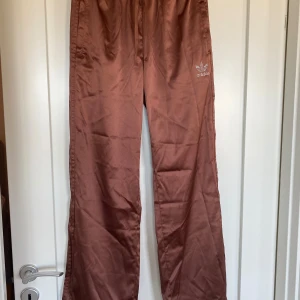 Bruna träningsbyxor från Adidas - Snygga bruna byxor från Adidas med en avslappnad passform. Byxorna har en elastisk midja och är dekorerade med Adidas-loggan i vita pärlor på sidan. Endast provade. Aldrig använda eller tvättade. Inga defekter. 