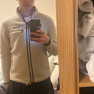 Beige fleecetröja från Patagonia - Säljer en mysig beige fleecejacka från Patagonia. Jackan har dragkedja framtill och praktiska fickor med dragkedjor. Perfekt för kyliga dagar och har en stilren design med mörkare kantdetaljer. Dock en liten defekt på vänster sida.
