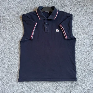 Moncler Piké - Säljer en stilren mörkblå pikétröja från Moncler med klassisk krage och knappar. Tröjan har röda och vita detaljer på kragen och ärmarna samt Monclers logga på bröstet. Perfekt för en avslappnad men ändå elegant look. Perfekt nu inför sommaren🤩Hör gärna av er vid frågor och funderingar! MVH Lavish