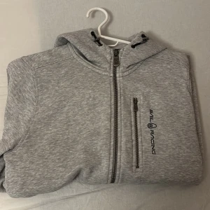 Grå hoodie från Sail Racing - Säljer en stilren grå hoodie från Sail Racing med dragkedja och justerbar huva. Passar dig som är 170-175 (S)