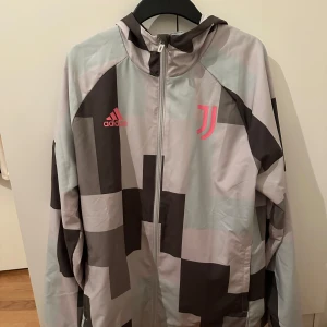 Grå och rosa vindjacka från Adidas - Säljer en grå och rosa vindjacka från Adidas med Juventus-logga. Jackan har ett unikt rutigt mönster och dragkedja framtill. Perfekt för sportiga aktiviteter eller som en stilren vardagsjacka.