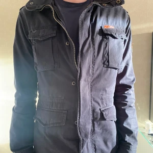 Marinblå jacka från Superdry - Säljer en marinblå jacka från Superdry med flera fickor och en Union Jack-detalj på ärmen. Jackan har dragkedja och knappar framtill samt en hög krage. Perfekt för en stilren look. S men kan Passa M