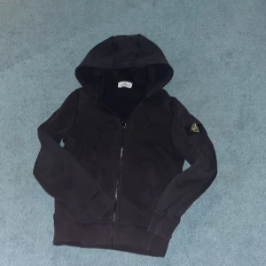 Svart hoodie från Stone Island - Snygg svart hoodie från Stone Island med dragkedja och huva. Den har en ikonisk logga på ärmen och är perfekt för en stilren look. Strorleken är i juinor skulle säga Xs, bara att höra av sig!!