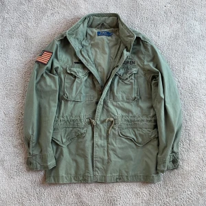 Ralph Lauren field jacket - Small - Säljer en  militärinspirerad jacka från Ralph Lauren med flera patchar, inklusive en amerikansk flagga och en flygdivisionsemblem. Jackan har dragkedja och knappar framtill samt flera fickor för praktisk förvaring. Perfekt för en cool och avslappnad stil. Passar Medium också. Ralph Lauren - skick 9/10 - storlek S 