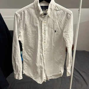 Vit Ralph Lauren skjorta  - Säljer en klassisk vit skjorta från Ralph Lauren i slim fit. Skjortan har långa ärmar och knappar framtill. Den är prydd med det ikoniska broderade logotypen på bröstet. Perfekt för en stilren look. Jag säljer den för att den är för liten. Den är ostyrkt som man ser med stryker såklart innan leverans! Under ärmarna så är det jätte lite gul som har sätt sig men men ser det ej ifall man inte kollar jätte noga så ingen man tänker på! Hör av dig ifall du är intresserad!!😊