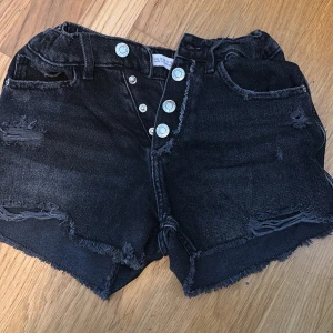 Svarta jeansshorts från Zara - Säljer ett par coola svarta jeansshorts från Zara med slitna detaljer och fransiga kanter. Shortsen har knappar framtill och klassiska femficksdesign. Perfekta för en avslappnad stil!