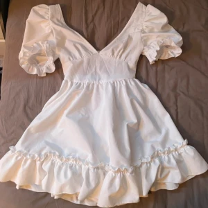 Vit klänning från SHEIN - Charmig vit klänning från SHEIN med puffiga kortärmar och volangdetaljer. Klänningen har en v-ringad design och en öppen rygg med elastisk detalj. Perfekt för sommarens varma dagar eller student.