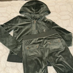 Grön velour hoodie och byxor från Juicy Couture - Säljer en stilren grön velour hoodie och matchande byxor från Juicy Couture. Hoodien har dragkedja och snörning i luvan, medan byxorna har elastisk midja och fickor med knappar. Perfekt för en avslappnad look!