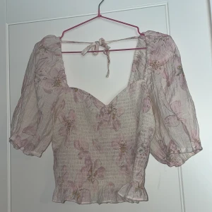 Blommig topp från H&M - Säljer en söt blommig topp från H&M med puffiga ärmar och knytning i ryggen. Toppen har en v-ringad design och är dekorerad med ett vackert blommönster i rosa och beige nyanser. Perfekt för en somrig look! 🌸