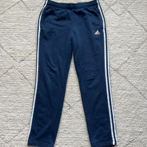 Mörkblå mjukisbyxor från Adidas - Säljer ett par mörkblå mjukisbyxor från Adidas med vita ränder längs sidorna. Byxorna har en elastisk midja och är perfekta för en avslappnad stil. Adidas-loggan är broderad på ena benet.