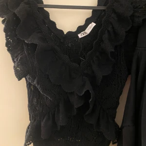Svart spetsblus från Zara som inte säljs längre - Säljer en elegant svart spetsblus från Zara med volangdetaljer och v-ringning. Perfekt för att ge en romantisk touch till din outfit. Blusen har en fin struktur och är tillverkad i ett lätt material.