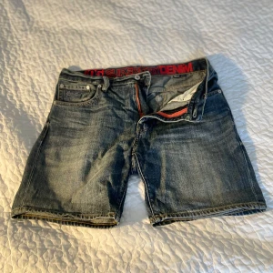 Jeansshorts från Superdry - Snygga jeansshorts från Superdry i klassisk blå denim. De har en normal passform och är perfekta för sommardagar. Shortsen har en dragkedja och knappar framtill samt flera fickor för praktisk användning.