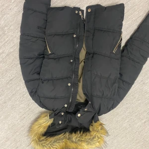 Svart dunjacka från Bershka - Säljer en svart dunjacka från Bershka med pälskantad huva och dragkedja. Jackan har långa ärmar och flera fickor med dragkedjor. Perfekt för kyliga dagar.