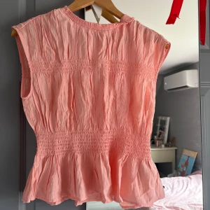 Rosa ärmlös blus - Söt corallrosa ärmlös blus med smockdetaljer och volang nedtill. Perfekt för varma dagar och ger en romantisk touch till din outfit.