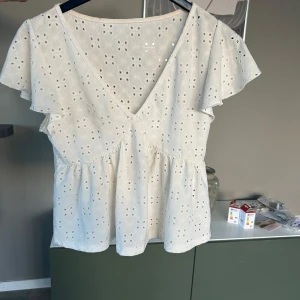 Vit blus med spetsdetaljer - Säljer en söt vit blus med v-ringning och spetsdetaljer. Blusen har korta ärmar med volang och ett fint broderat mönster. Perfekt för en somrig look!